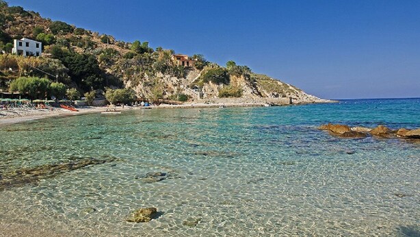 A coastal beach with clear shallow waters, rocky outcrops, and buildings on a hillside.