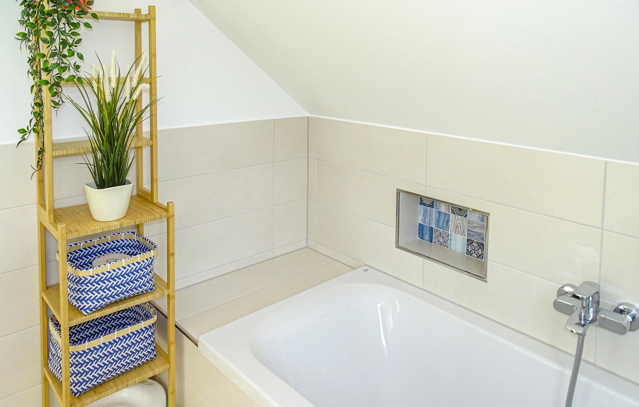 A bathroom with a wooden shelf holding a potted plant and blue patterned baskets, a tiled shower area, and a white bathtub.