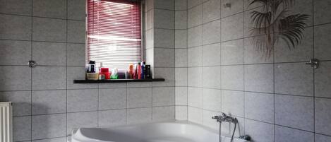 Ein Badezimmer mit Eckbadewanne, einem Fenster mit Jalousien und einem Ablagefach für Pflegeprodukte.