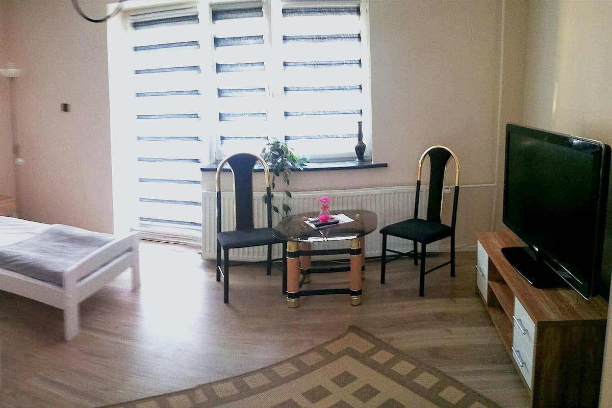 A small, well-lit room with a single bed, a chair, a small table, and a TV.