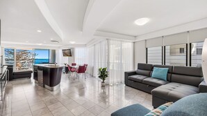 A modern living room with a grey sofa, a dining area with red chairs, and a kitchen with a central island. - Superb location for this level 4  two bedroom apartment, swim between the flags just accross the roa (Mooloolaba)