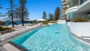A swimming pool with clear blue water, surrounded by trees and a building with balconies. - Superb location for this level 4  two bedroom apartment, swim between the flags just accross the roa (Mooloolaba)