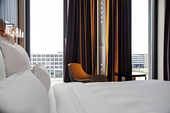 A hotel room with a bed, a chair, a small table, and a view of a building through large windows.
