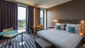 A hotel room with a large bed, a desk, a chair, and a view of the cityscape. - Steigenberger Airport Hotel Berlin (Schoenefeld)