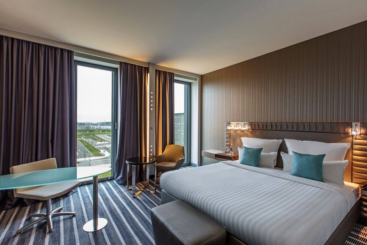 A hotel room with a large bed, a desk, a chair, and a view of the cityscape.