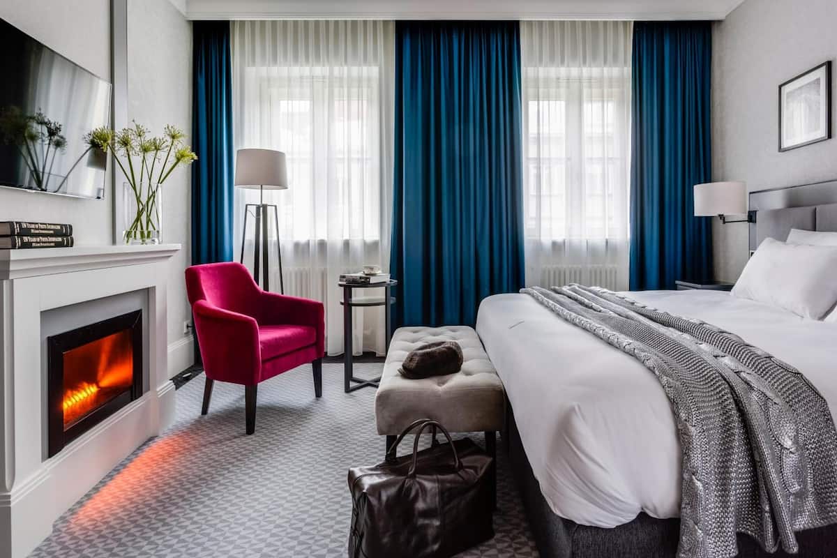 Hotellihuone, jossa on takka, sänky, punainen nojatuoli, pieni pöytä ja yöpöytä.