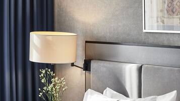 Ett hotellrum med en säng, vita kuddar, ett nattduksbord med en lampa, en vas med blommor och två inramade tavlor på väggen.