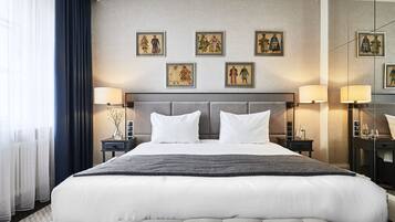 Una cama grande con cabecera gris, ropa de cama blanca y una manta gris. Hay dos mesitas de noche con lámparas y un espejo. La habitación tiene un ventanal con cortinas translúcidas y un cortinero azul oscuro.