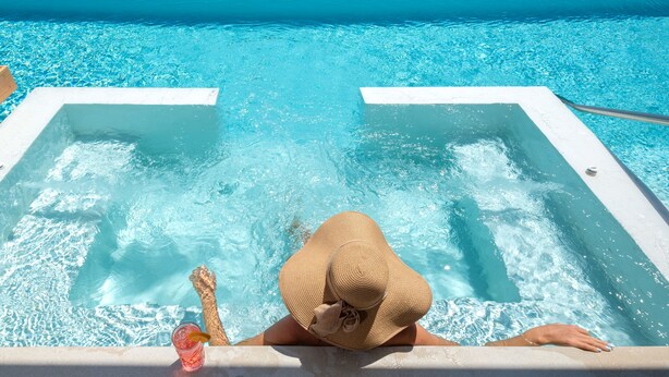 Una piscina con agua azul cristalina, una persona relajándose en un jacuzzi y vistas al océano y a una carretera que conduce a un edificio.