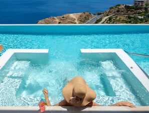 A pool with clear blue water, a person relaxing in a hot tub, and a view of the ocean and a road leading to a building. - Villa Lucrezia, Minimalist Design & Spa Retreat (Heraklion)