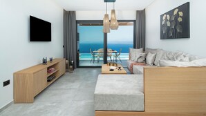 Una sala de estar moderna con un ventanal grande, un televisor de pantalla plana, un mueble bajo de madera, un sofá y una zona de comedor con vista al océano.