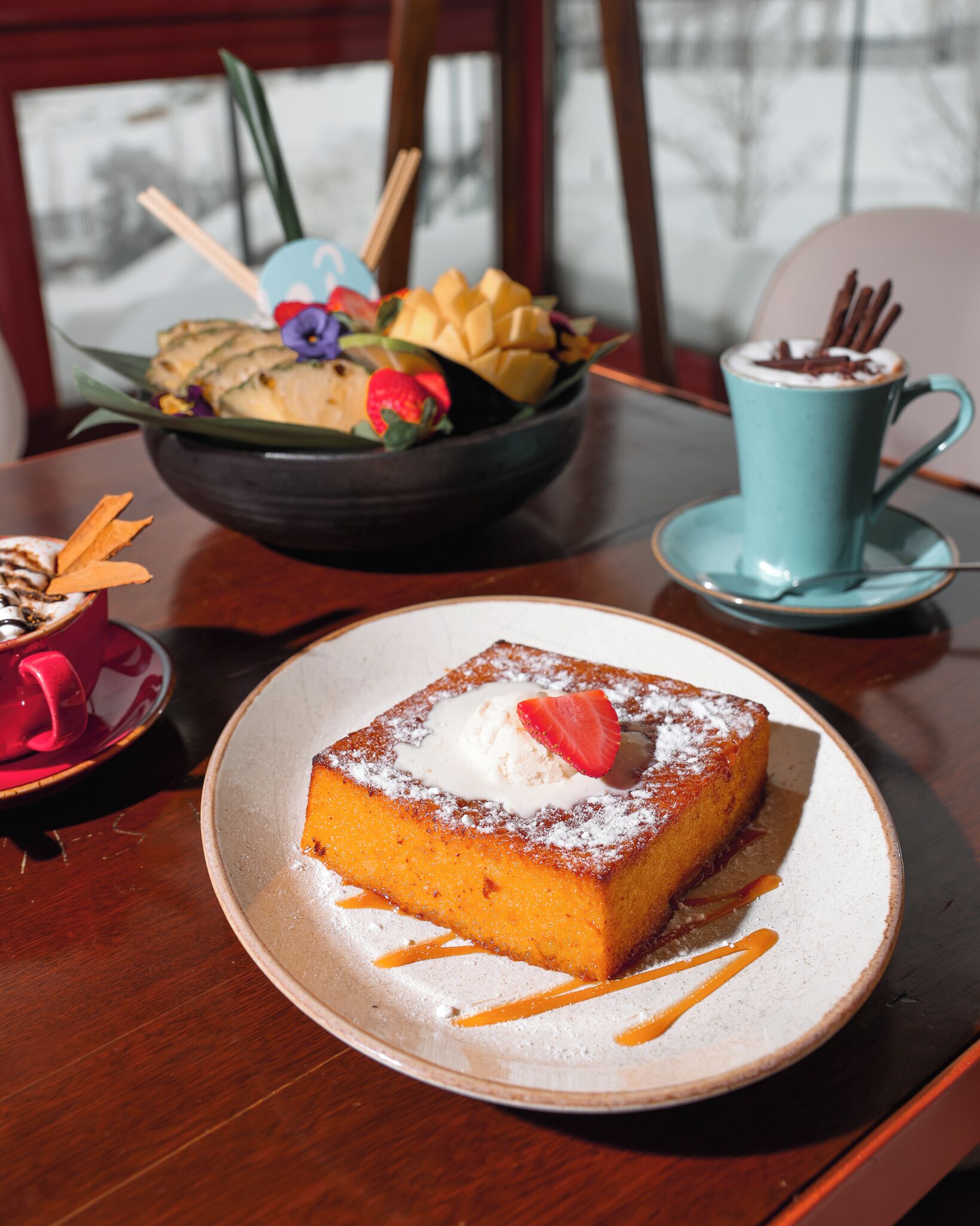 A dessert plate with a piece of cake topped with whipped cream and a strawberry, a bowl of fruit, and a blue cup with a spoon.