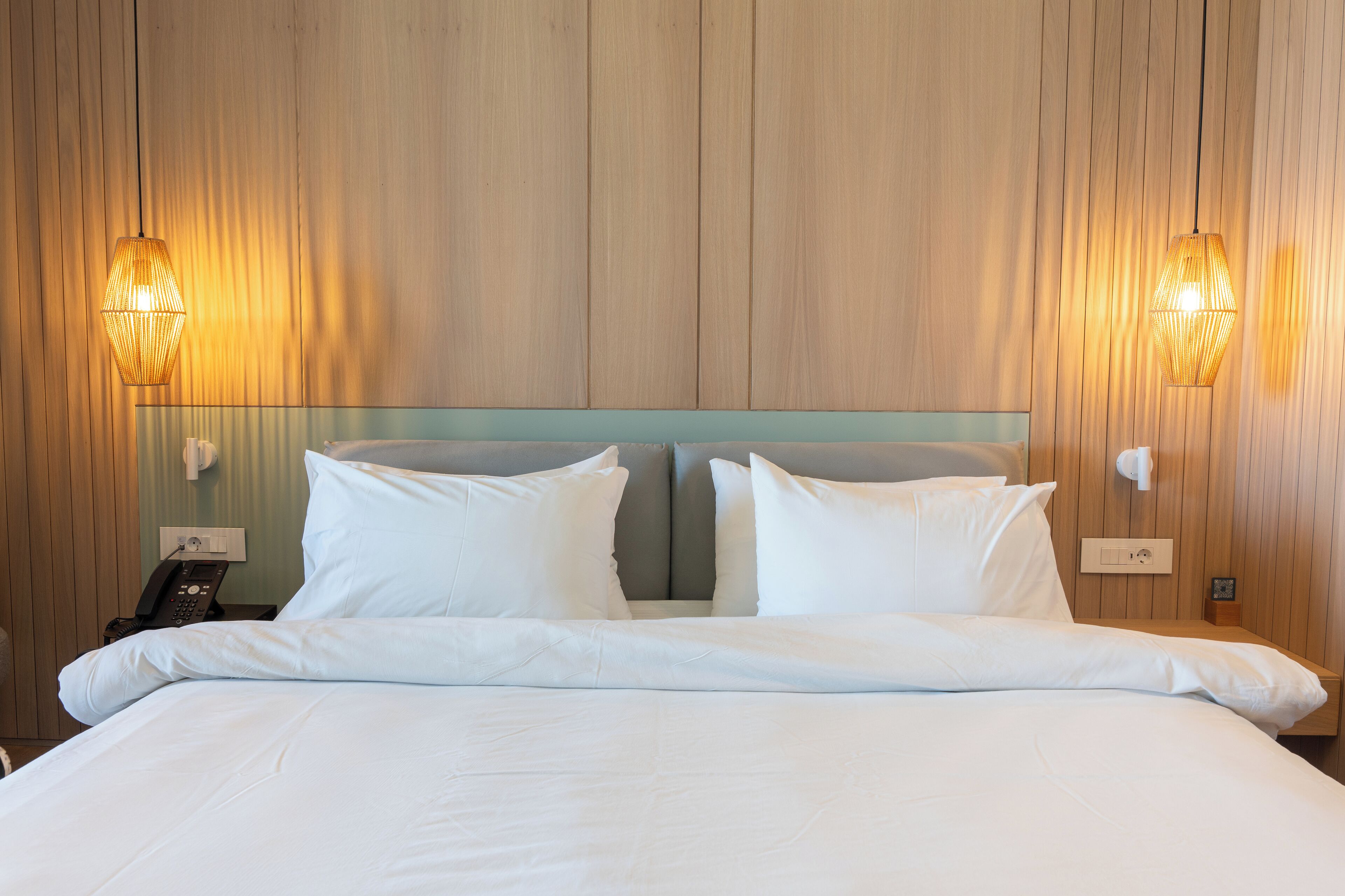 A neatly made bed with white linens and pillows, a wooden headboard, and two wall-mounted lamps.
