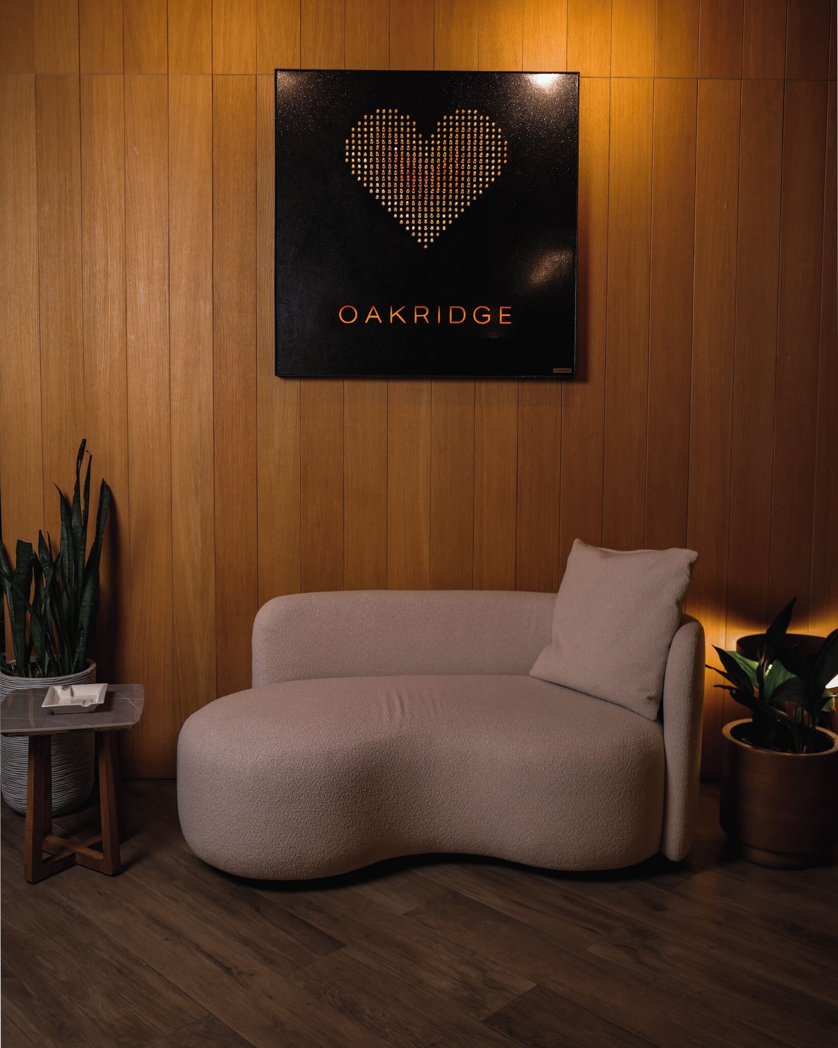 Een kamer met een gebogen bank, een kleine tafel met een plant, en een hartvormig bordje aan de muur met de tekst 'OAKRIDGE'.