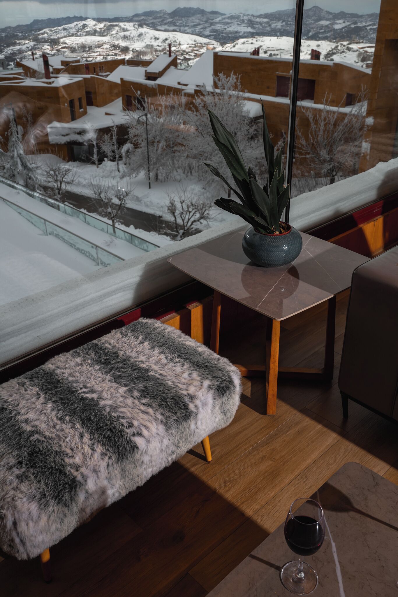 Un interior moderno con vistas a un paisaje nevado, una mesa de madera, un banco y una copa de vino tinto.