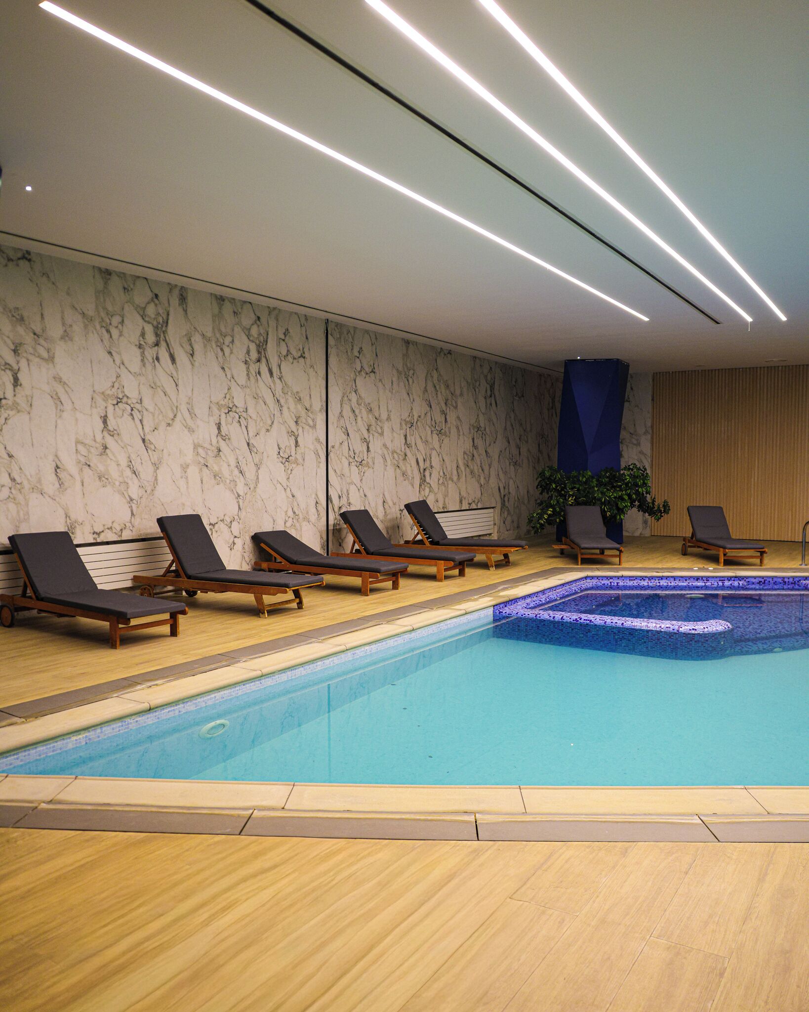 A swimming pool with lounge chairs, a marble wall, and a wooden floor.