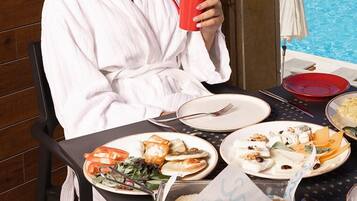 A person in a white bathrobe sitting at a table with plates of food and a red mug, overlooking a pool with a mountainous view.