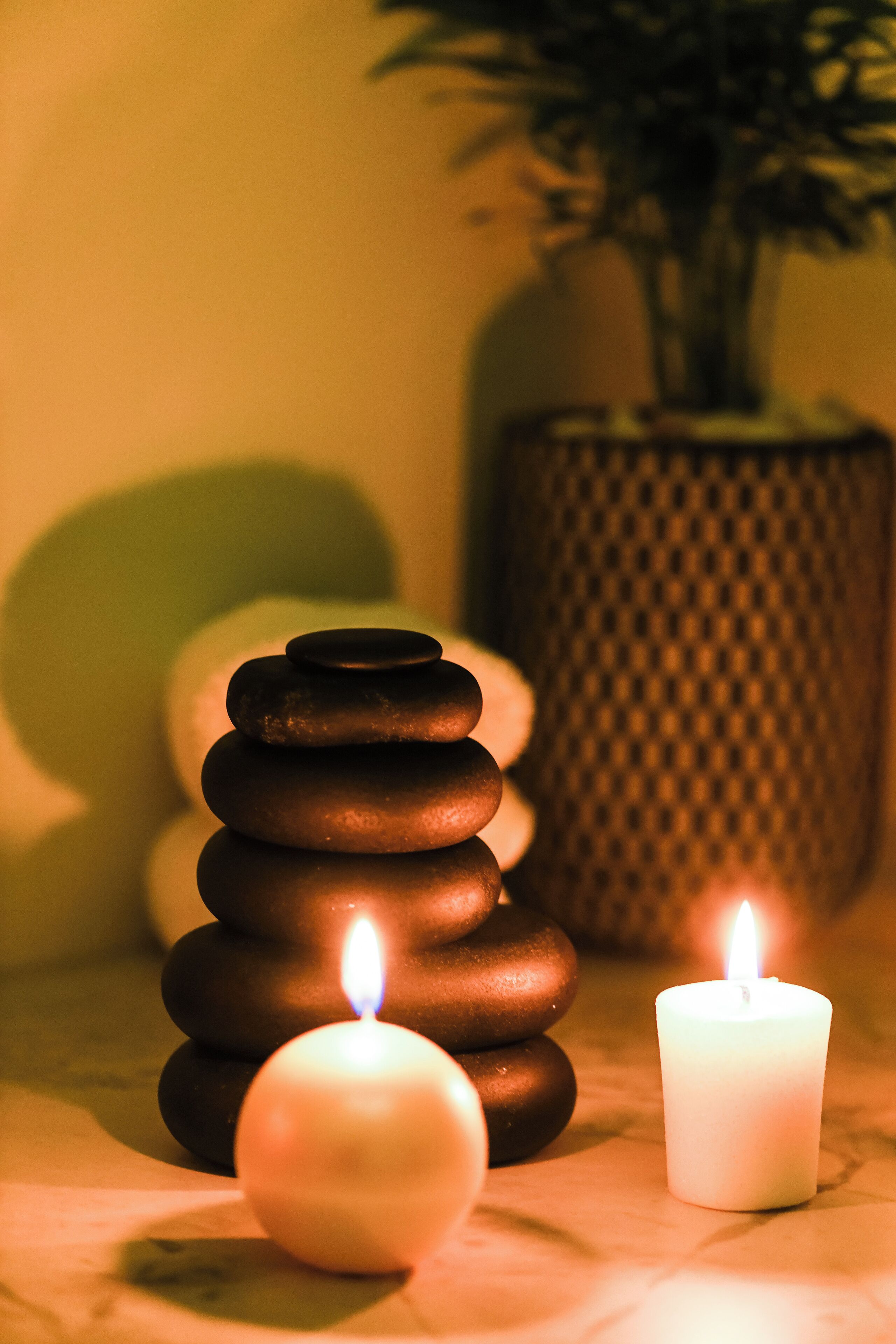 A stack of smooth stones and two lit candles on a table, with a textured vase and a potted plant in the background.