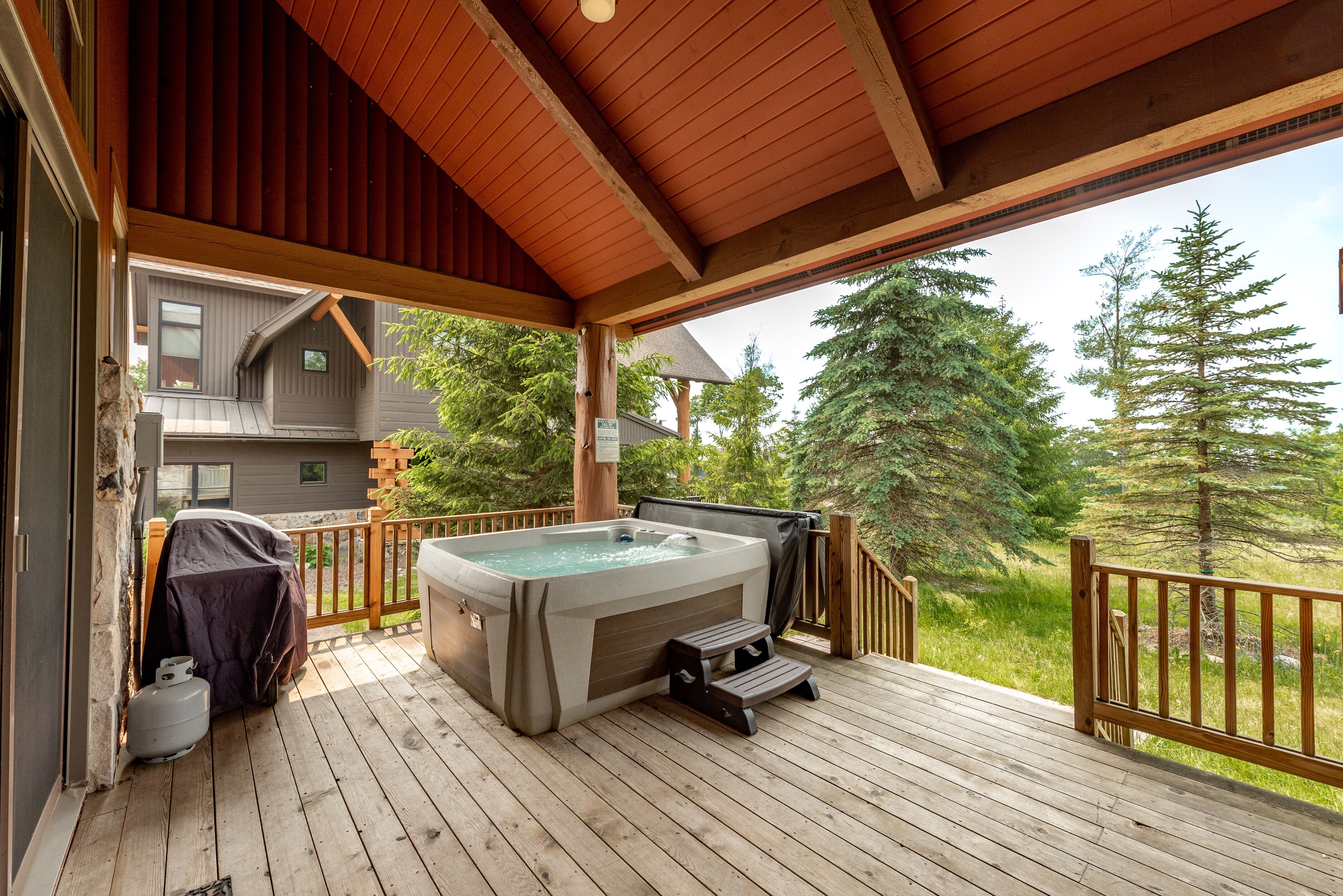 Una terraza de madera con jacuzzi, parrilla cubierta y un escalón que lleva a una casa.