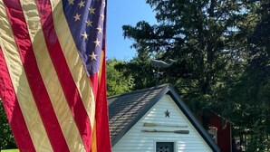 Ein kleines, weißes Haus mit einer dunkelblauen Tür, einem Stern an der Vorderseite und einem Fahnenmast mit einer amerikanischen Flagge.