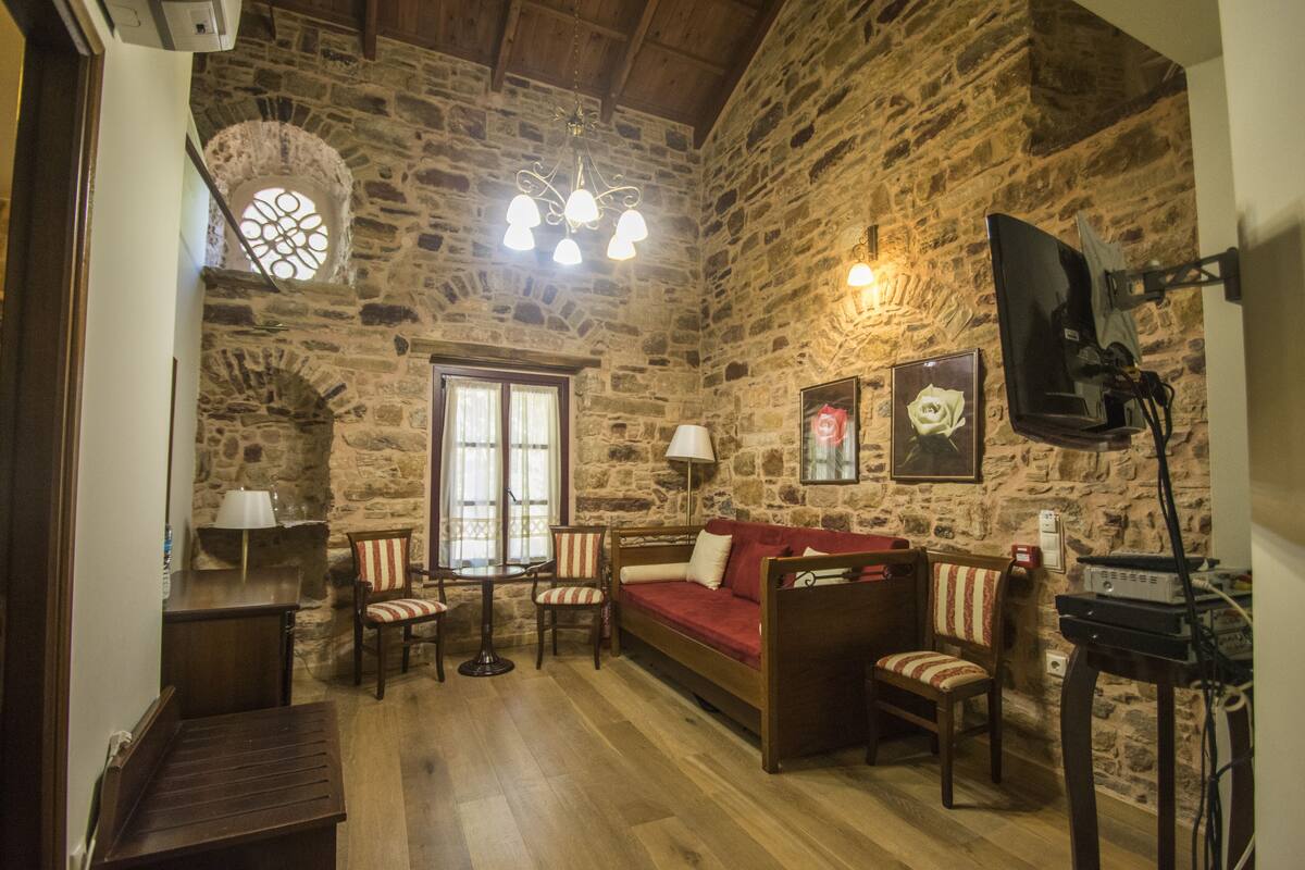 A stone-walled room with a wooden floor, a red sofa, striped chairs, a small table, and a ceiling light fixture.