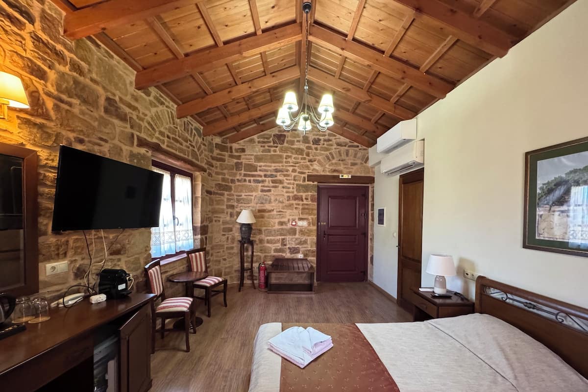 A cozy room with stone walls, wooden ceiling beams, a flat-screen TV, a desk with a computer, a bed with a brown and beige bedspread, and a chandelier.
