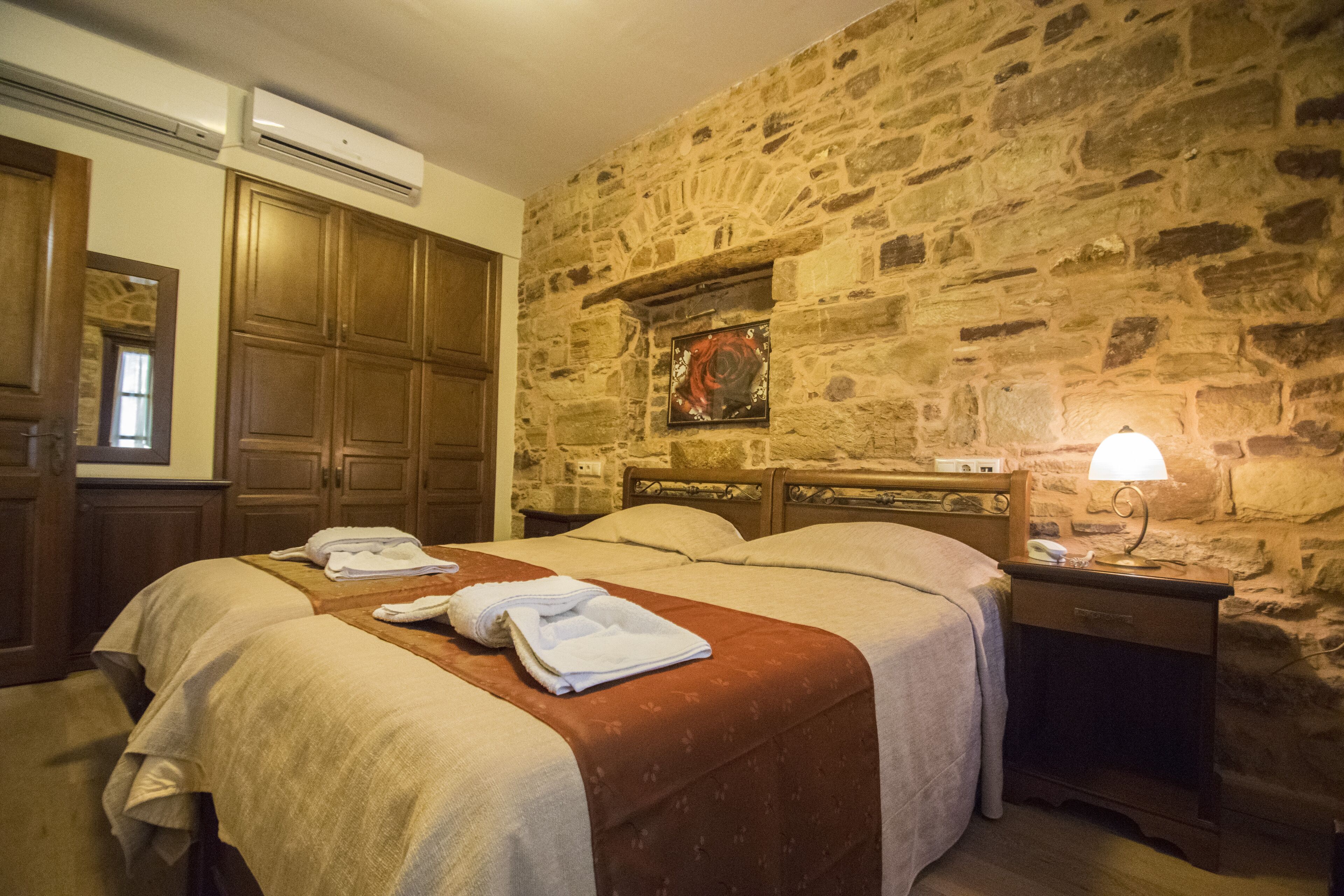 Quarto de hotel com parede de pedra, guarda-roupa de madeira, cama com colcha vermelha e mesa de cabeceira com abajur.