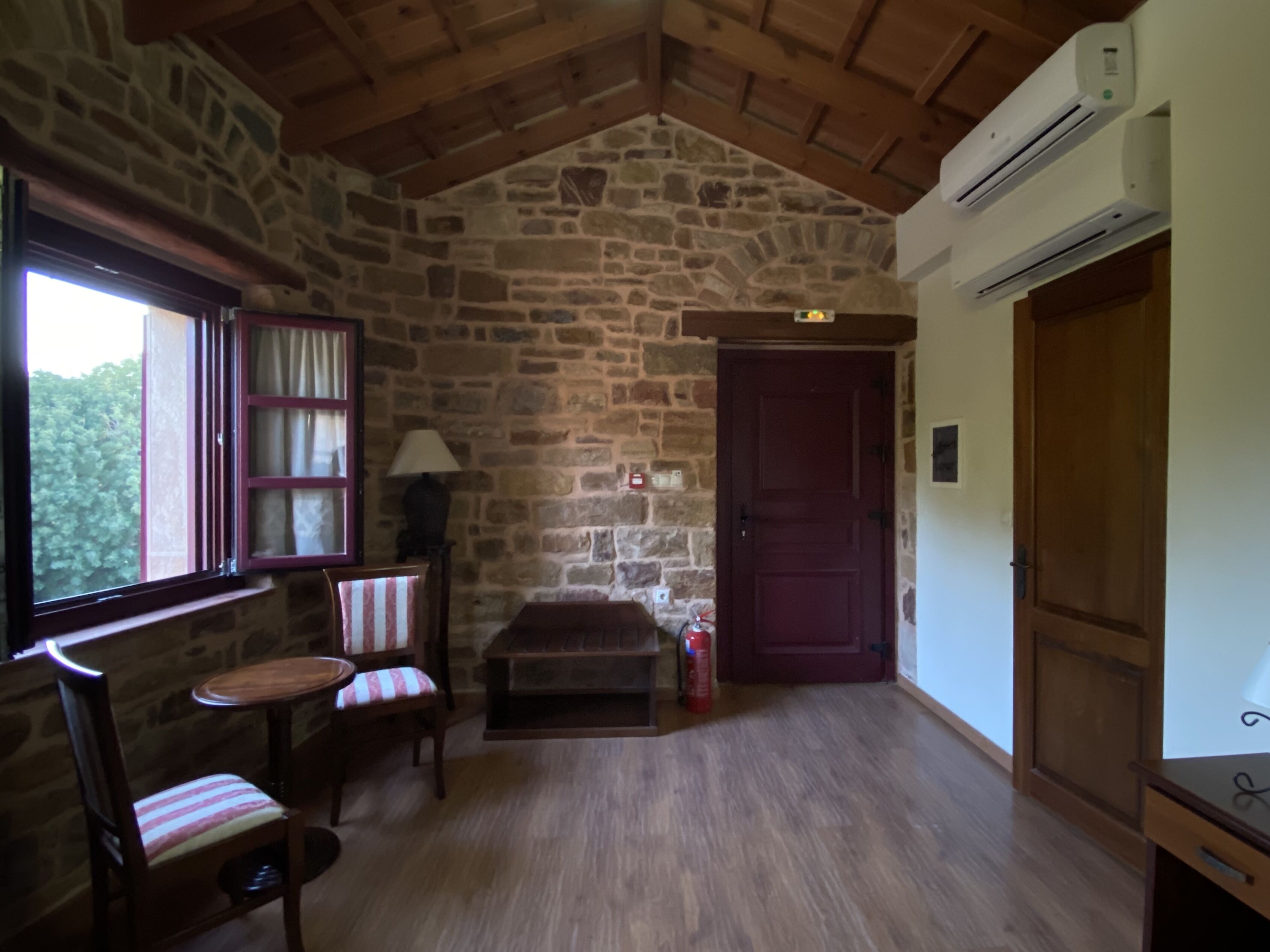A cozy room with stone walls, a wooden ceiling, a window with curtains, a chair with a striped cushion, a small table, and a door with a fire extinguisher.