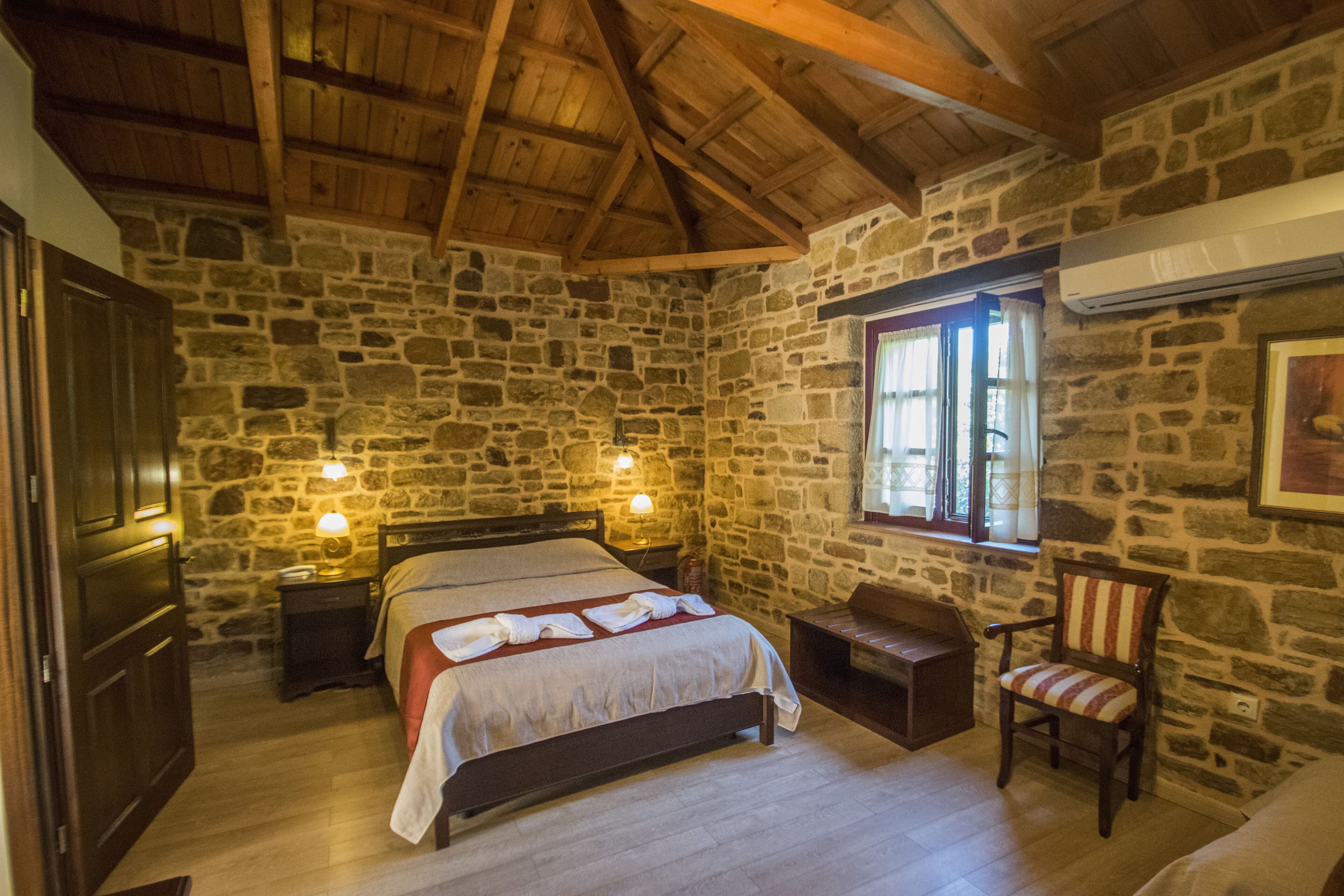 Ein Schlafzimmer mit Steinwänden und Holzboden, ein Bett mit roter Decke, zwei Nachttischlampen, ein Sessel und ein Fenster mit Vorhängen.