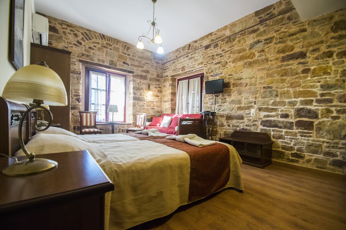 A room with a stone wall, a bed with a red headboard, a wooden nightstand, a lamp, and a television mounted on the wall.