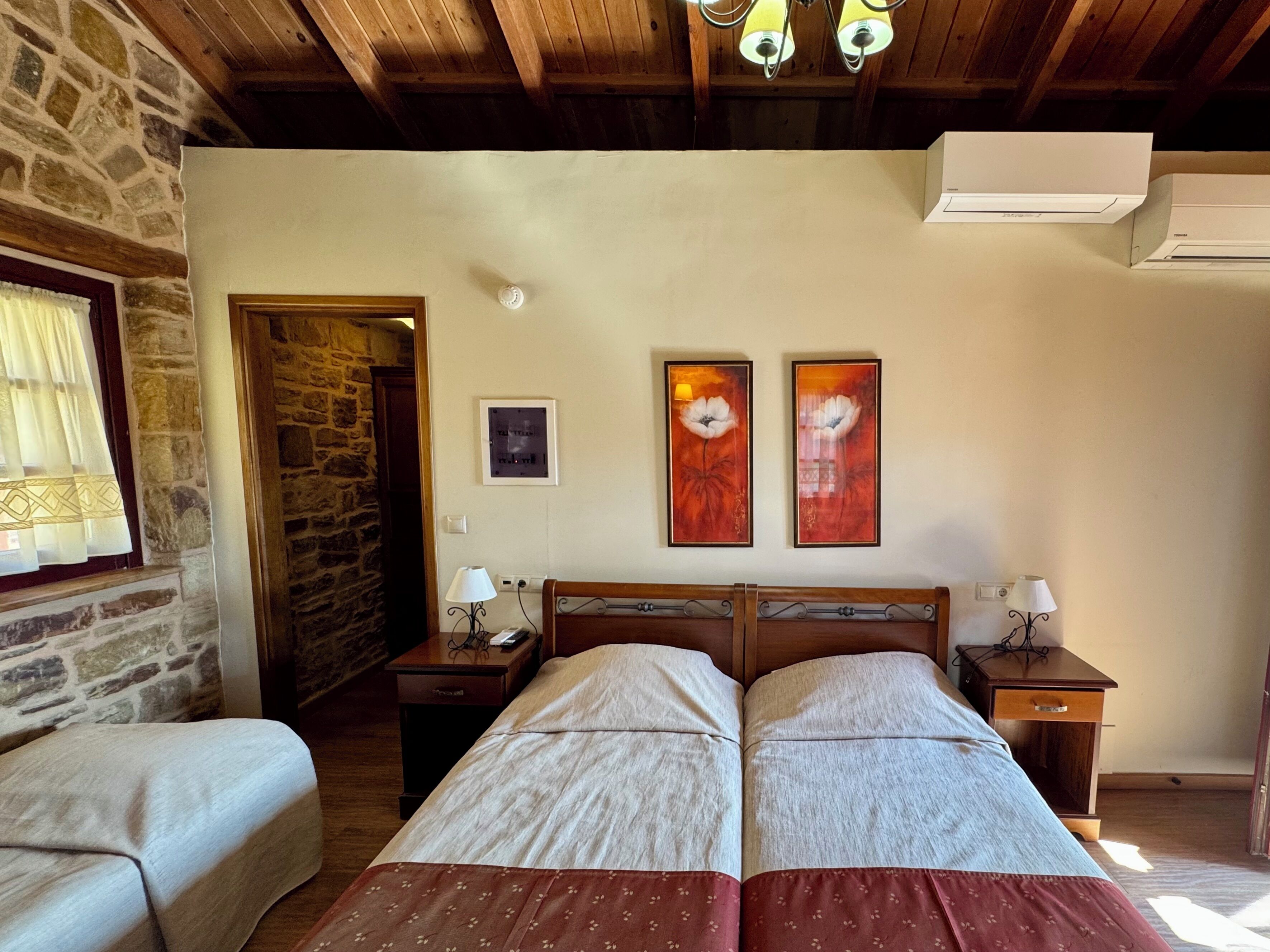 A room with a stone wall, wooden ceiling, two beds with red and white bedding, two bedside tables with lamps, and two framed artworks on the wall.