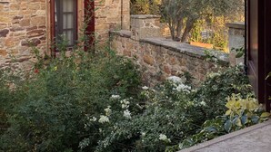 Un edificio de piedra con tejado de tejas rojas, una puerta de madera y un ventanal pequeño. Un jardín con diversas plantas y flores se encuentra frente al edificio.