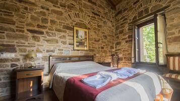 Un dormitorio con paredes de piedra, una cama, una mesita de noche, una lámpara y una ventana con cortinas.