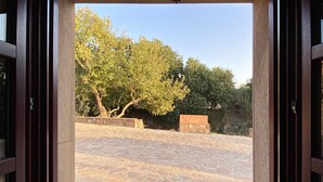 Vista a través de una ventana con marco de madera de un patio empedrado con árboles y un muro de piedra.