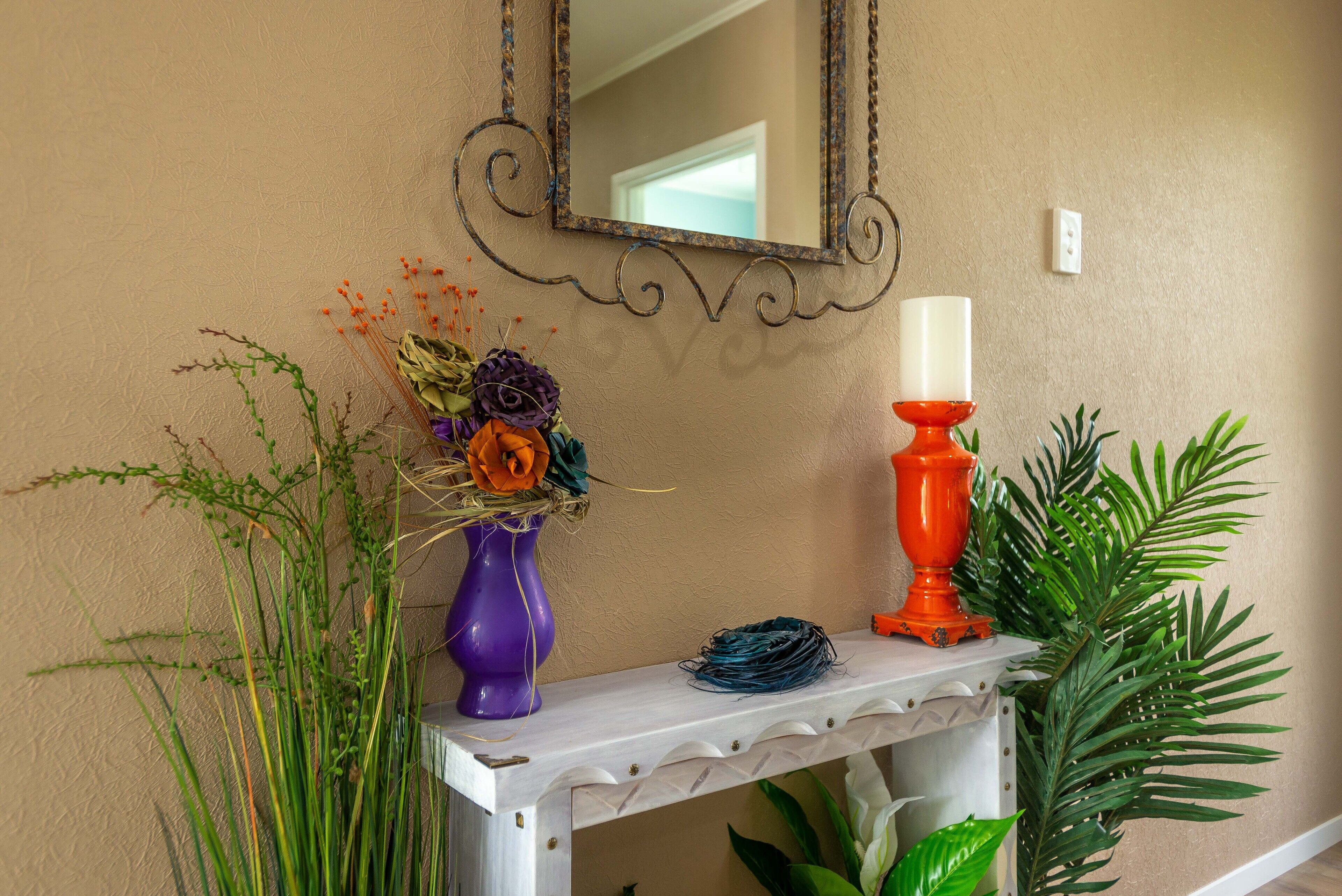 Ein weißer Konsolentisch mit Spiegel, einer violetten Vase mit Blumen, einem orangefarbenen Kerzenhalter und grünen Pflanzen.