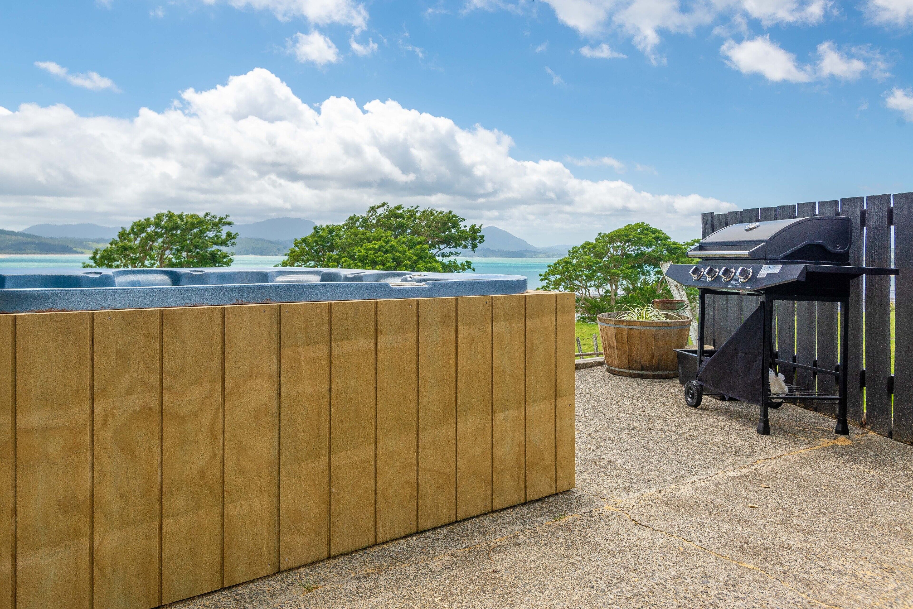 Une terrasse en bois avec un jacuzzi, un barbecue et une vue sur l’océan et les montagnes.