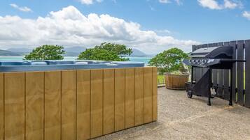 Une terrasse en bois avec un jacuzzi, un barbecue et une vue sur l’océan et les montagnes.