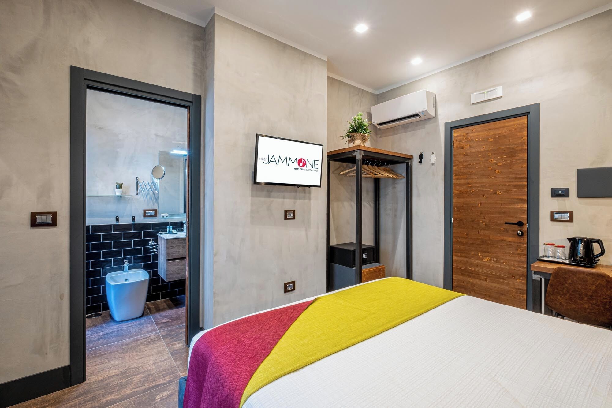 Camera d'albergo moderna con un letto, bagno con lavandino e specchio, armadio e televisore che mostra il nome dell'hotel.