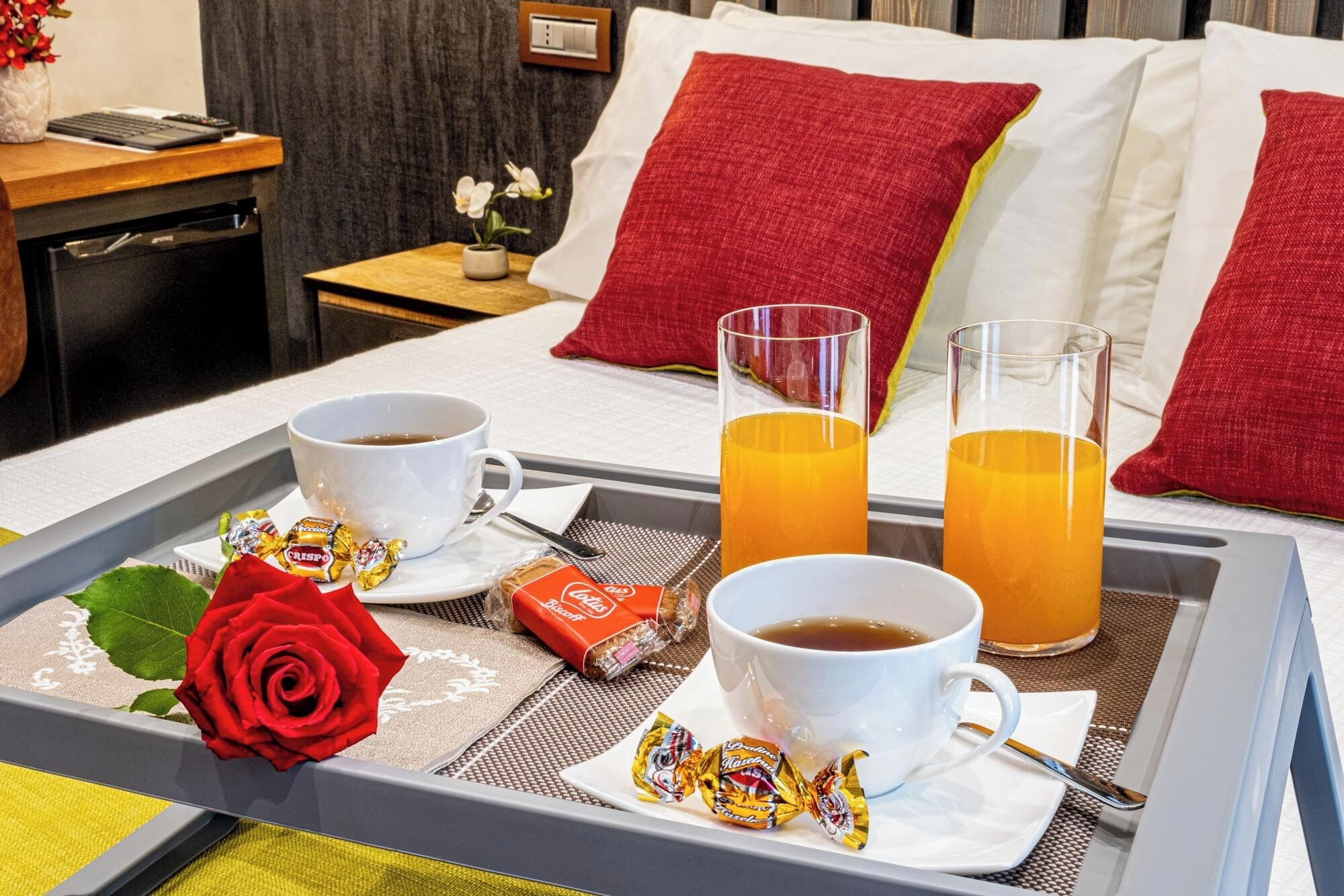 A hotel room with a bed, a bedside table with a lamp, a tray with breakfast items including two cups of coffee, two glasses of orange juice, a red rose, and some chocolates.