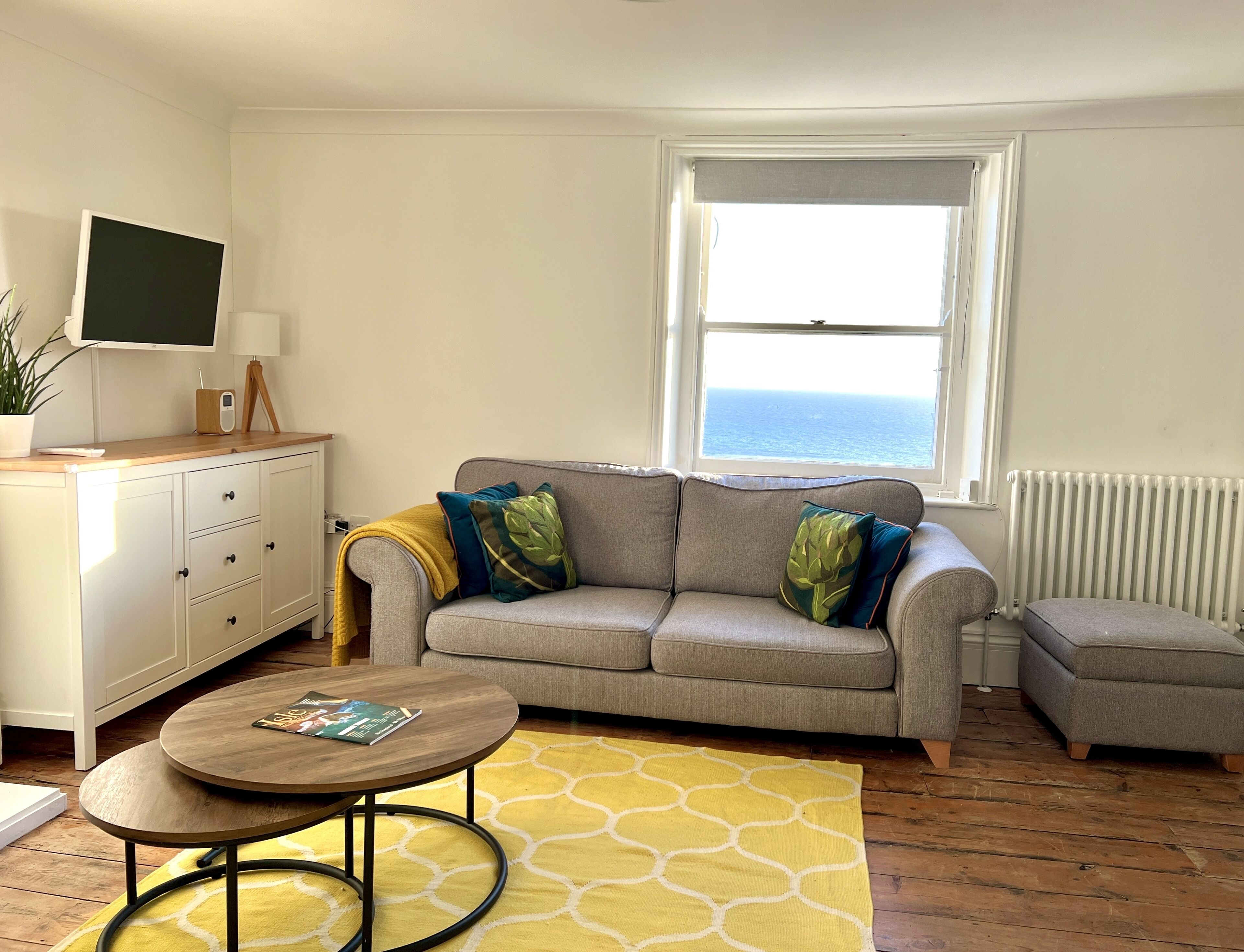 Un salón con sofá gris, una mesita de madera, alfombra amarilla, un mueble blanco, televisor sobre un soporte y ventana con vista al mar.