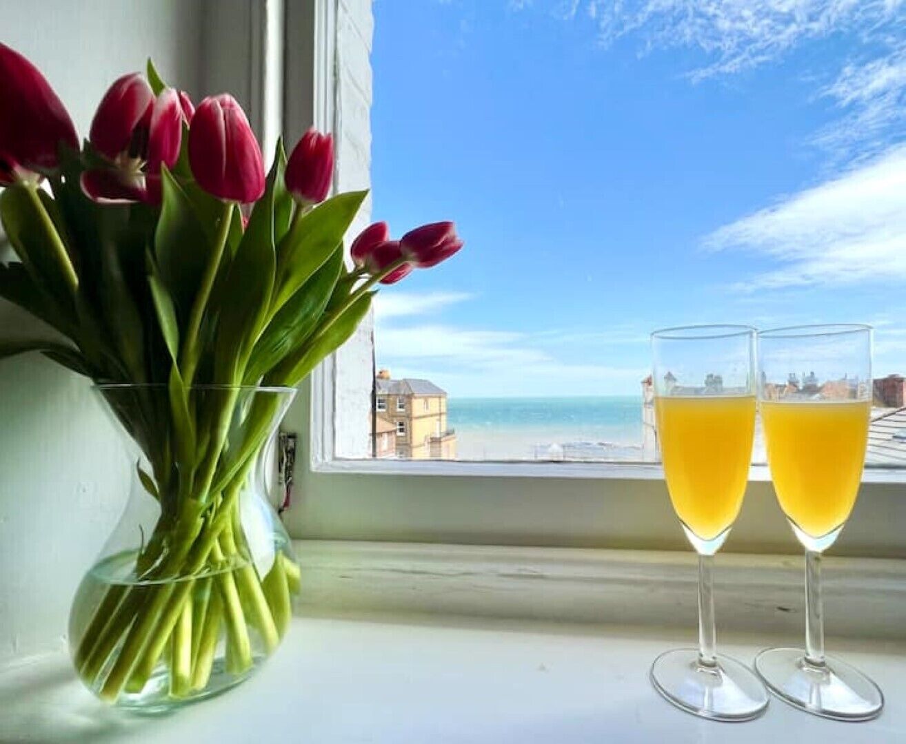 Vista da janela para a praia, um vaso com tulipas e dois copos de suco de laranja.