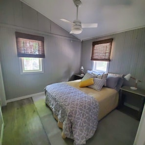 Un dormitorio con una cama, dos mesitas de noche, un ventilador de techo y dos ventanas con persianas.