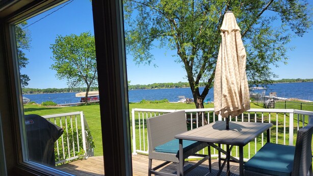 Une terrasse avec une table et des chaises, donnant sur un lac où l’on peut voir des bateaux et une zone herbeuse.