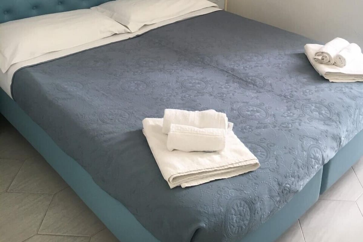 A neatly made bed with a blue headboard and white linens, a small bedside table, and a wall-mounted light fixture.