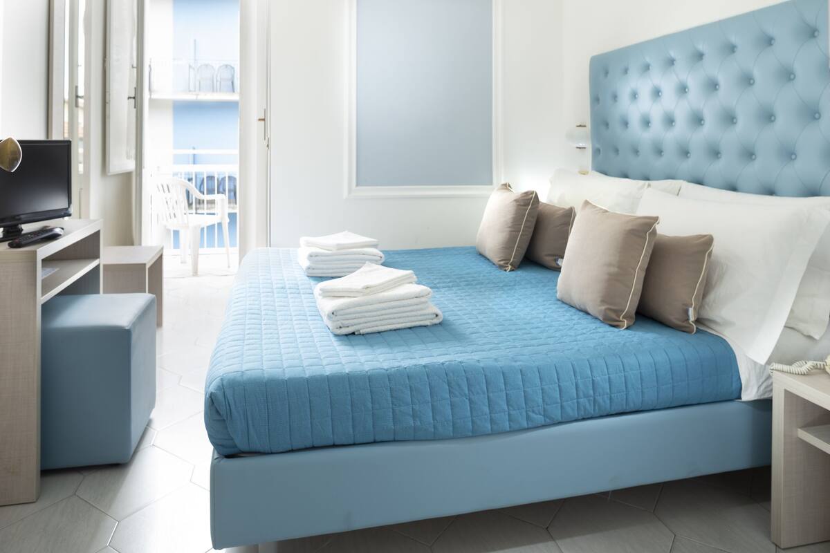 A modern hotel room with a large bed, a television, and a balcony with white chairs.