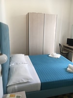 Una camera d'albergo con un letto, un armadio, una scrivania con una sedia e una lampada.