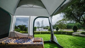A tent with a bed inside, a grassy area outside, and a view of trees and a body of water. - Campistaan Badlapur (Ambernath)