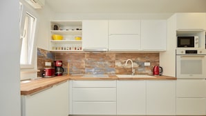 Une cuisine moderne avec des meubles blancs, un plan de travail en bois, un évier, une bouilloire rouge, une machine à café, un four à micro-ondes et un four traditionnel.