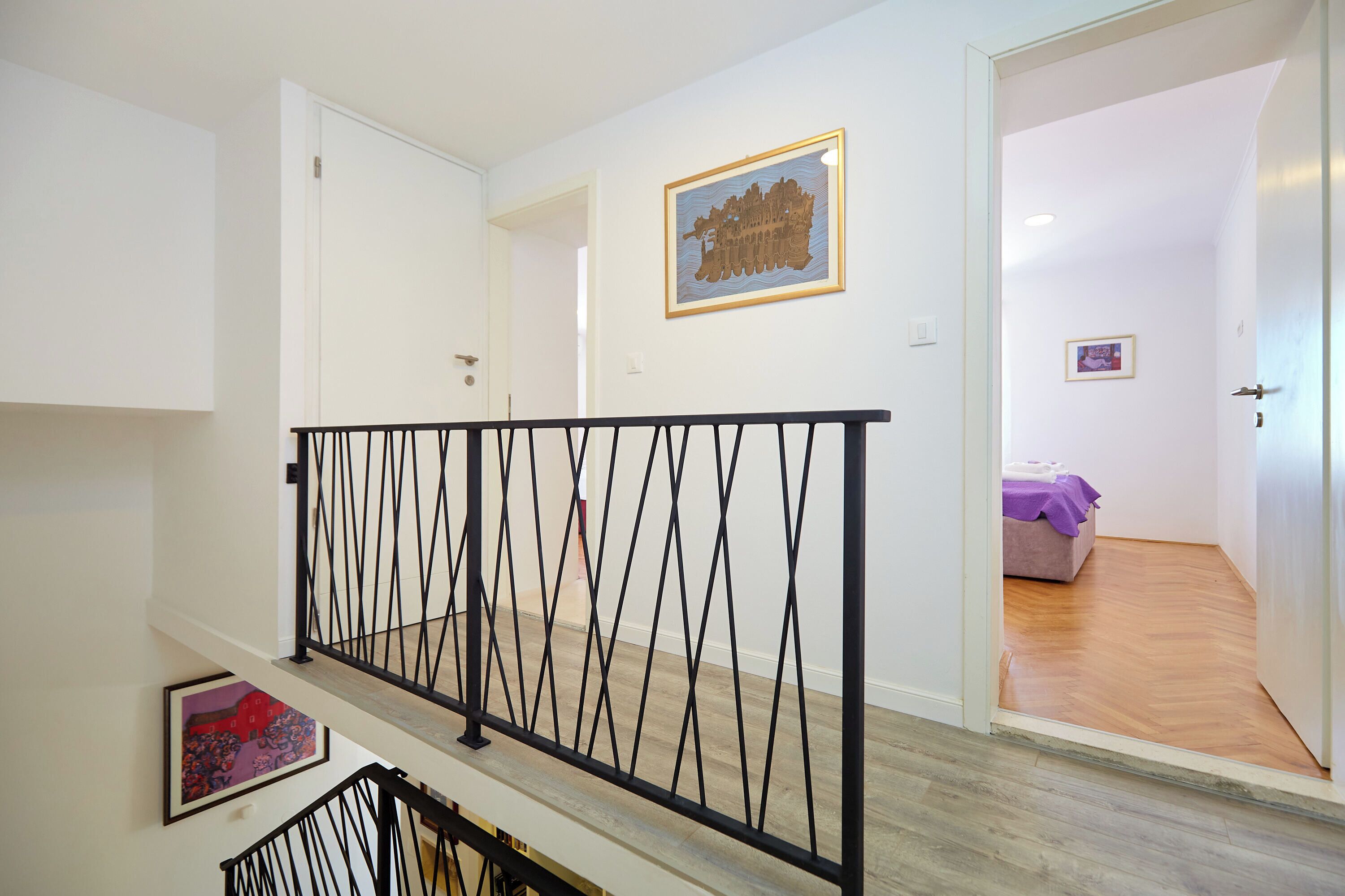 A staircase with a black metal railing, a wooden floor, and a door leading to a room with a purple bedspread.