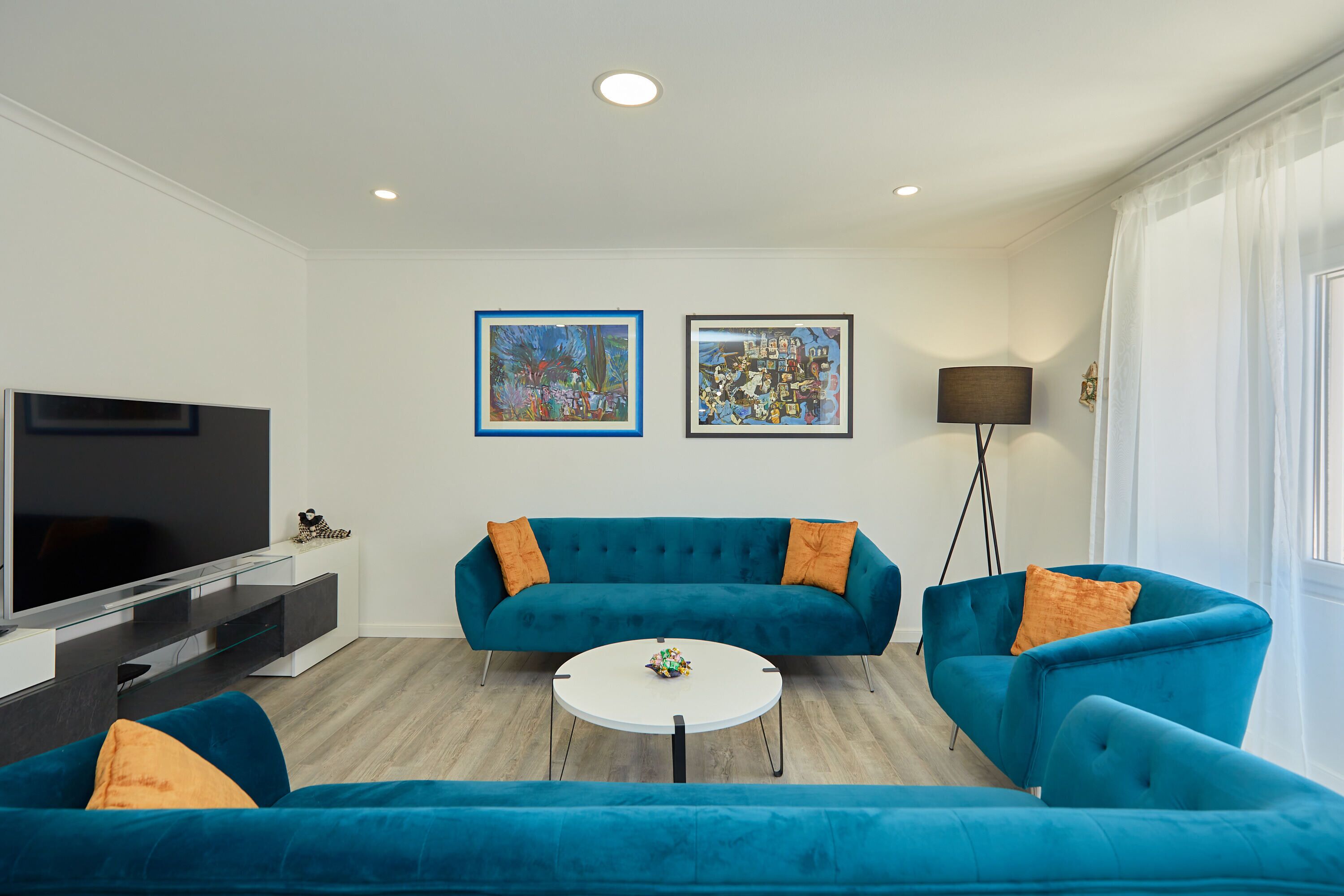 A modern living room with a teal sofa set, a flat-screen TV, and two framed artworks on the wall.